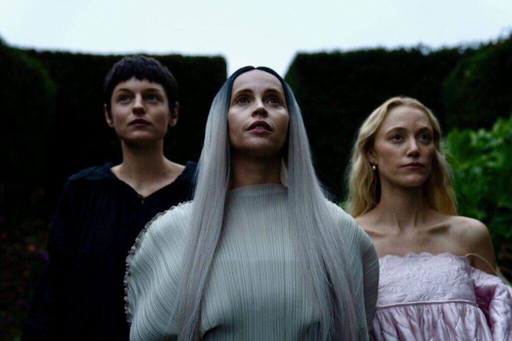 Three women stand closely together outdoors. The central woman has long silver hair and wears a pale flowing dress. The woman on the left has short dark hair, and the woman on the right has long blonde hair and wears an off-shoulder dress.