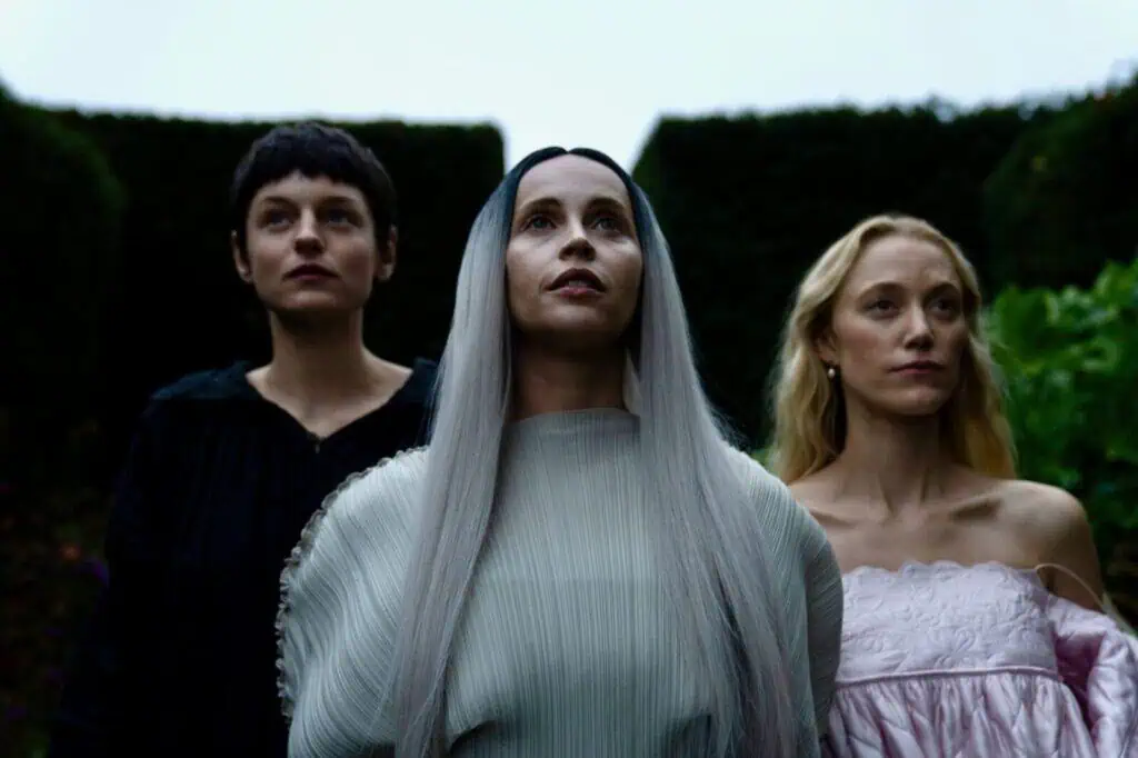 Three women stand closely together outdoors. The central woman has long silver hair and wears a pale flowing dress. The woman on the left has short dark hair, and the woman on the right has long blonde hair and wears an off-shoulder dress. Still from "It’s an Absurd World" directed by Julia Jackman.