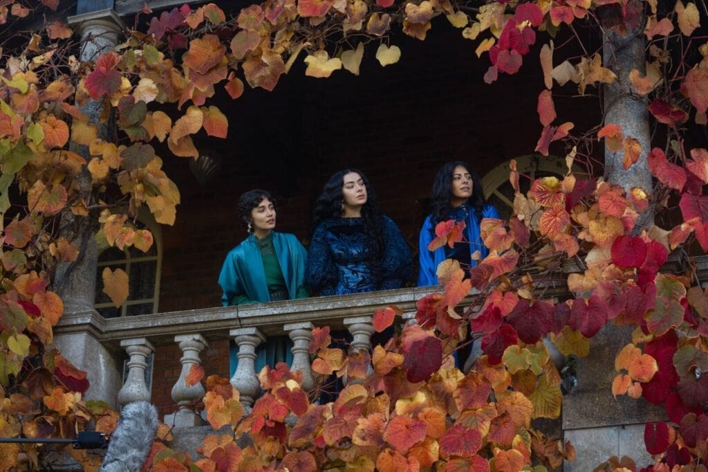 Three women in colorful clothing stand on a stone balcony surrounded by vibrant autumn leaves in shades of orange, yellow, and red, looking thoughtfully into the distance.