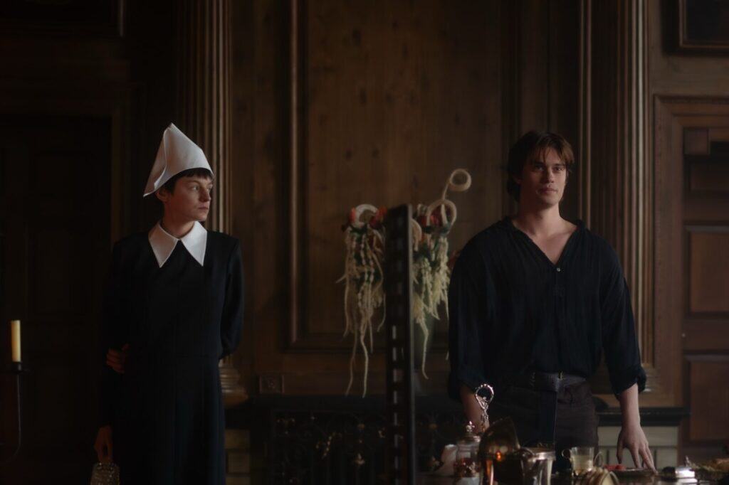 Two people stand in a dimly lit, wood-paneled room. One wears a black dress with a white collar and a tall white bonnet, while the other wears a black shirt with rolled sleeves. Ornate decorations and objects are on a table in front of them.