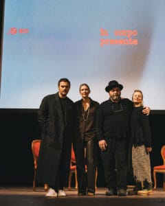 Sabato De Sarno, Jacopo Benass on a stage in front of a screen displaying the words in corpo presente and the IED logo. Three red chairs are visible behind them.