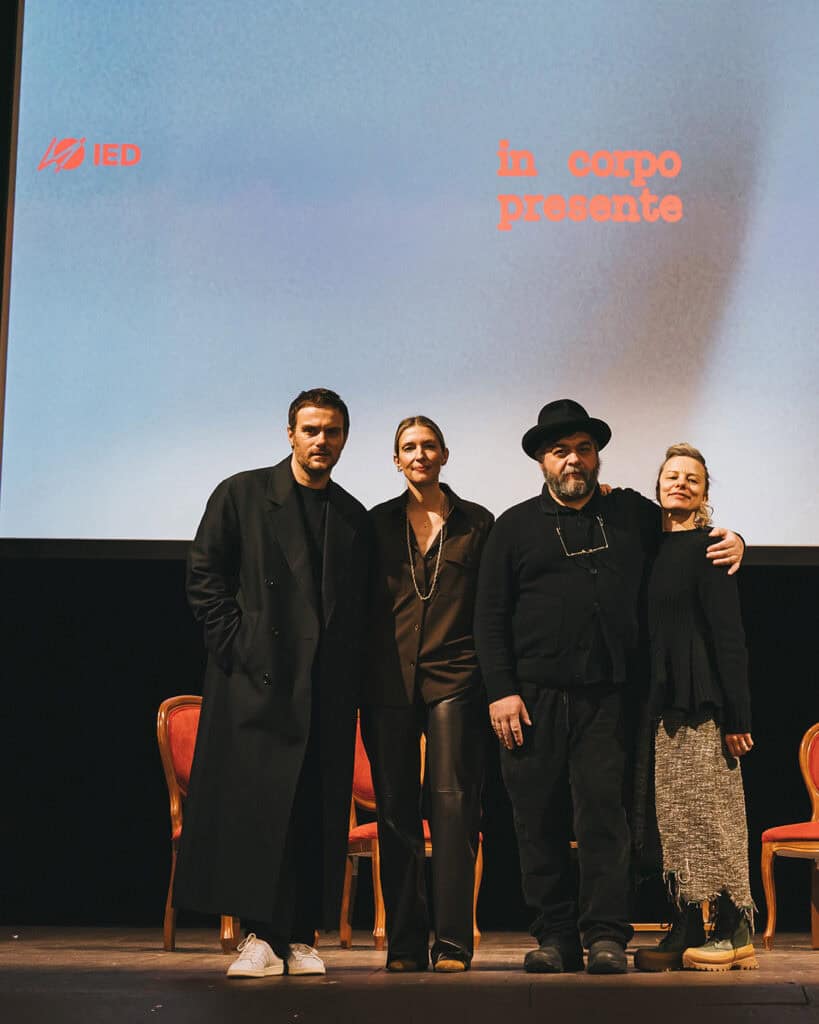 Sabato De Sarno, Jacopo Benass on a stage in front of a screen displaying the words in corpo presente and the IED logo. Three red chairs are visible behind them.