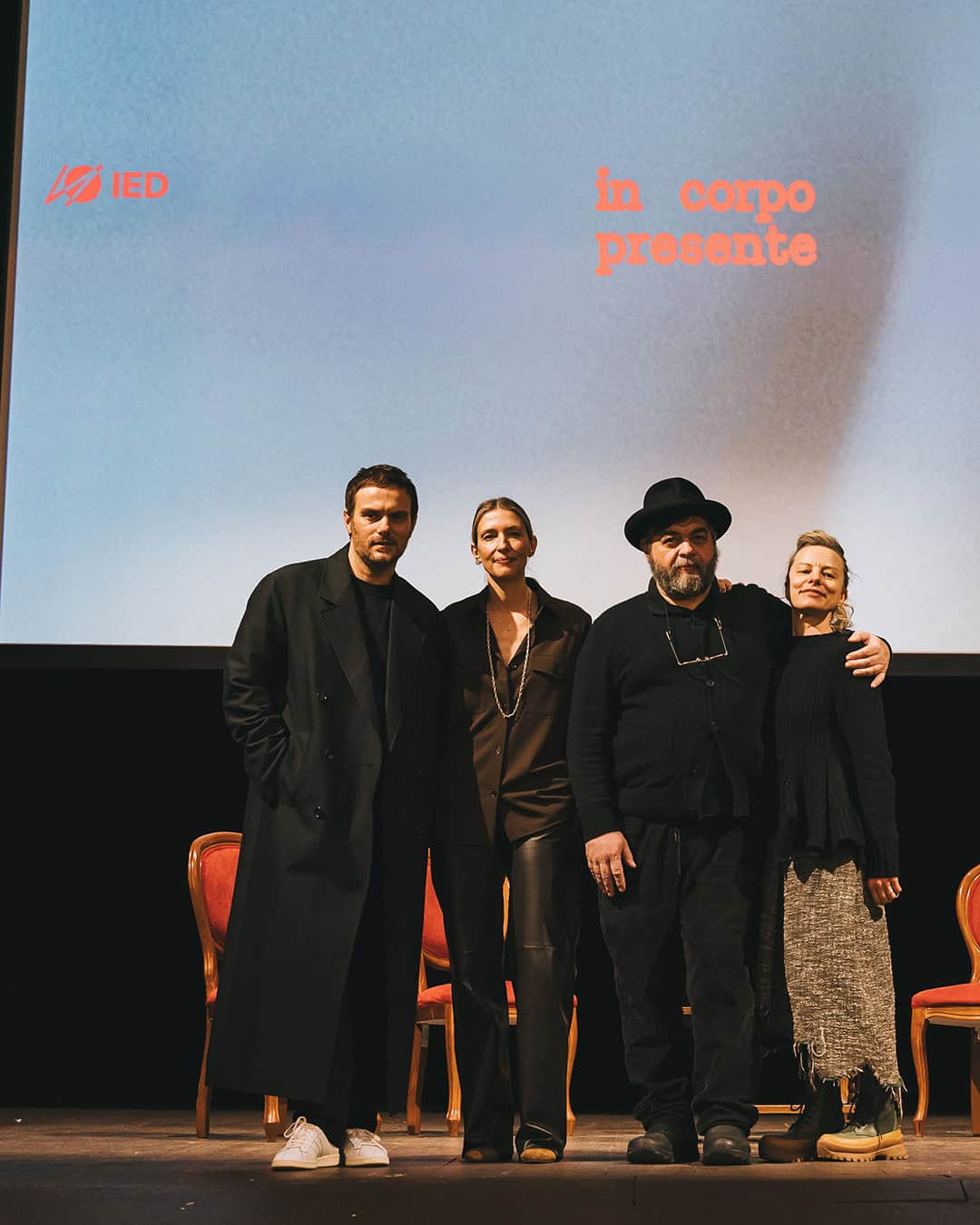 Sabato De Sarno, Jacopo Benass on a stage in front of a screen displaying the words in corpo presente and the IED logo. Three red chairs are visible behind them.