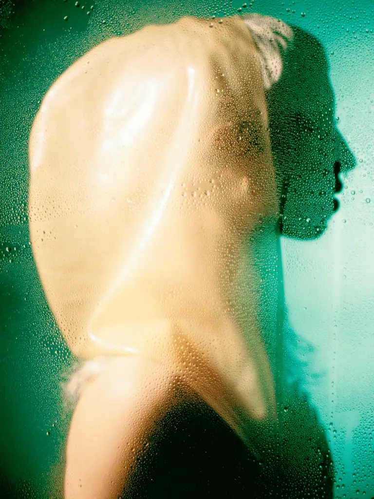 A person wearing a translucent hood stands behind foggy, wet glass, with dramatic lighting casting their shadowed profile against a teal background, evoking the mysterious allure of London’s nightlife.
