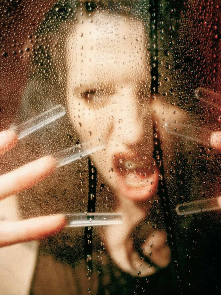 A person with long nails and an intense expression presses their face and hands against a wet, foggy glass surface covered in water droplets.