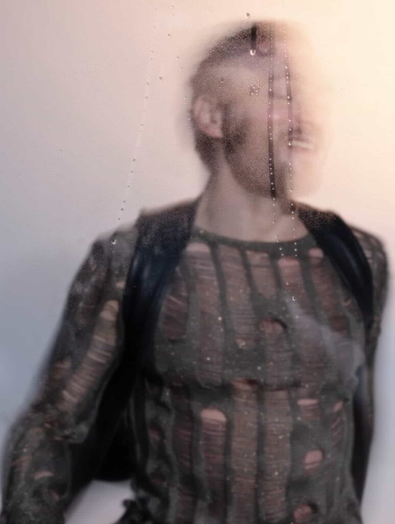 A person with a blurred face stands behind a foggy glass pane with water droplets, wearing a distressed, see-through top and dark suspenders or backpack straps.