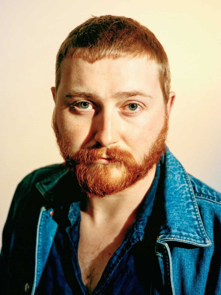 A person with short red hair and a full red beard looks directly at the camera. They are wearing a blue denim jacket over a dark shirt, with a neutral, softly lit background.