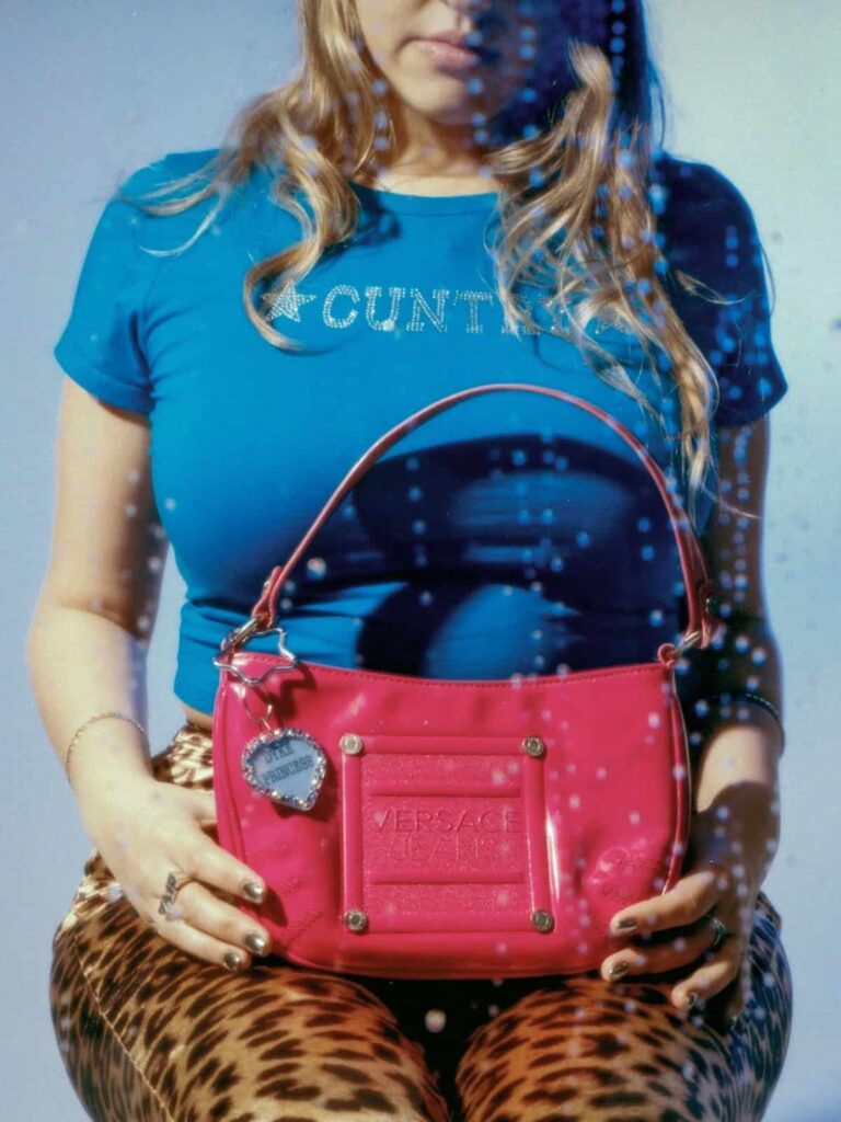 A person wearing a blue rhinestone-studded shirt and leopard print pants holds a bright pink Versace handbag. The photo has blue spots, possibly water droplets, and the persons face is partly out of frame.