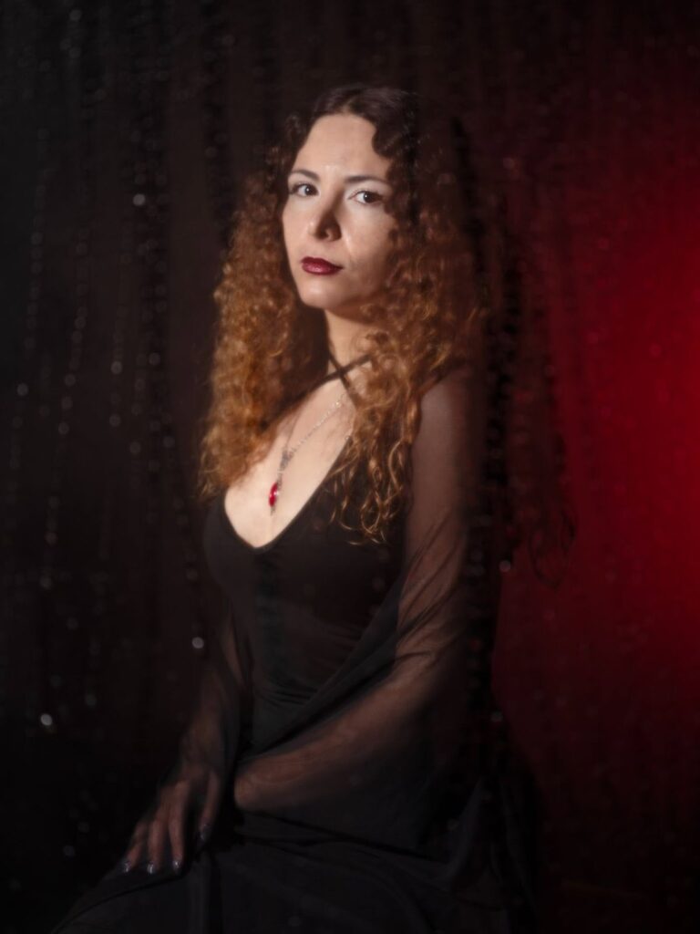 A woman with long curly hair wearing a black dress and a red pendant necklace sits against a dark background with a sheer black curtain and red lighting on one side.