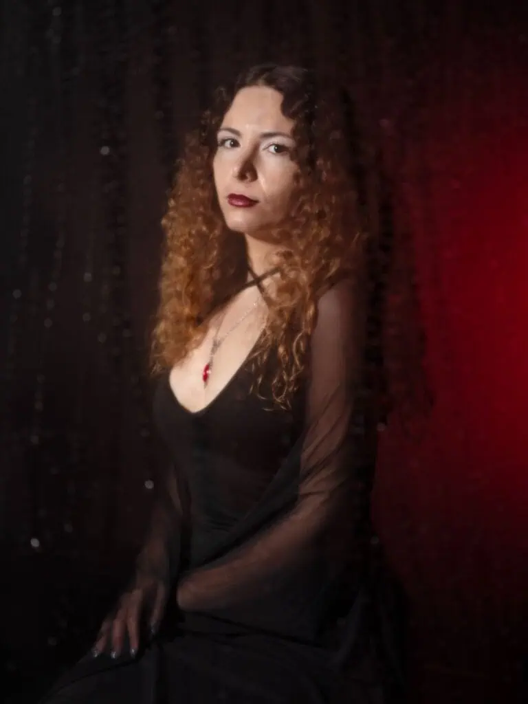 A woman with long curly hair wearing a black dress and a red pendant necklace sits against a dark background with a sheer black curtain and red lighting on one side.