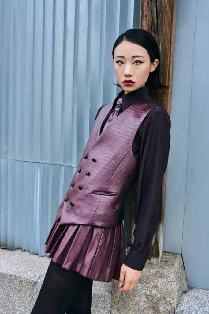 A woman with short black hair, wearing a shiny purple FRANCESCA COTTONE double-breasted vest over a pleated skirt and dark shirt, stands against a metal and wood wall, looking confidently at the camera.