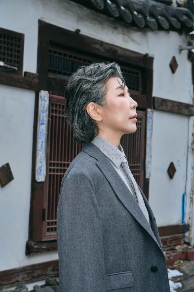 A person with short gray hair in a gray FRANCESCA COTTONE suit and polka dot shirt stands in profile outdoors, with a traditional-style building featuring wooden windows and blue calligraphy in the background.