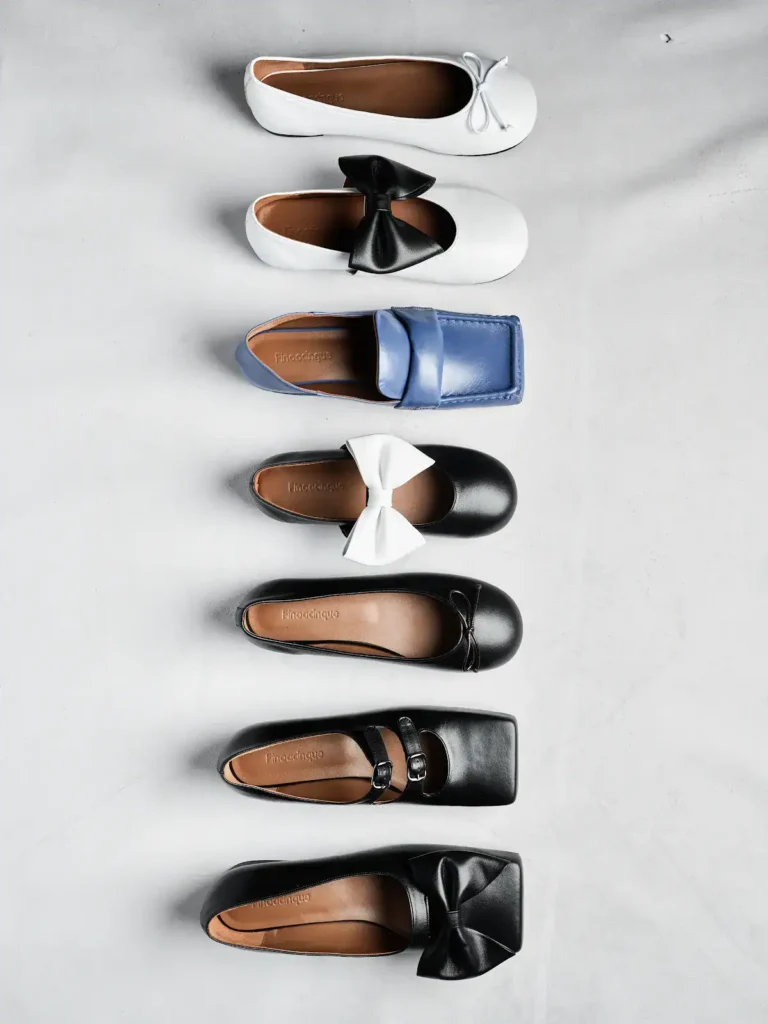 A vertical row of six womens flat Finoacinque shoes in various styles and colors, including white, blue, and black, each with different bow or strap details, displayed on a light gray surface.