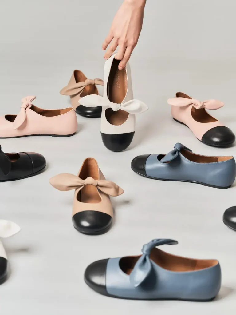 A hand holds a beige flat Finoacinque shoe with a large bow and black toe cap. Several similar shoes in beige, pink, and blue with black toe caps and bows are scattered on a light gray surface.