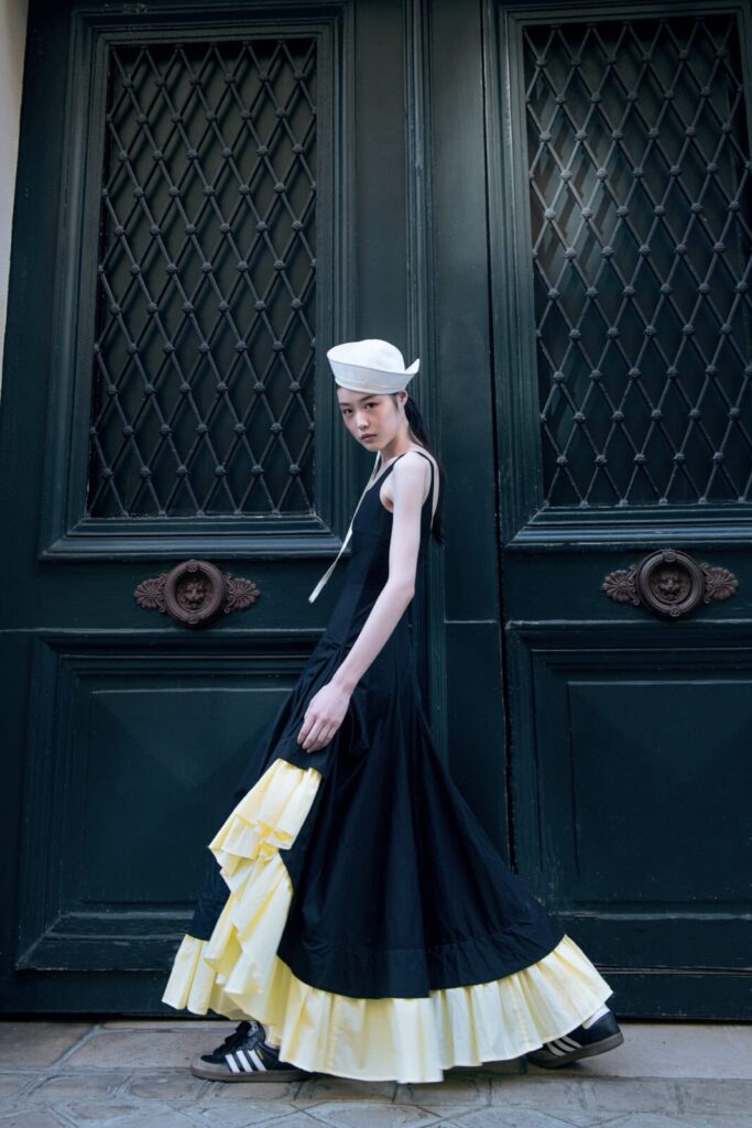 A person wearing a long black Denisa Rad dress with yellow ruffles, black sneakers, and a white hat stands in front of large dark green double doors with decorative metal grilles.