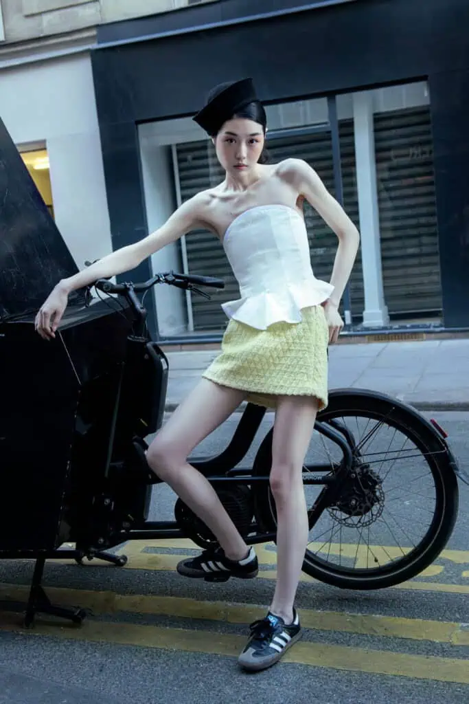 A woman in a strapless Denisa Rad white top, light yellow mini skirt, black beret, and black sneakers leans against a black bicycle on a city street, posing confidently with one hand on her hip.