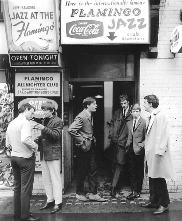 Five young men in 1960s attire stand talking outside the entrance of the Flamingo Jazz Club in London, under signs advertising jazz and allnighter events. The scene is candid and black and white.