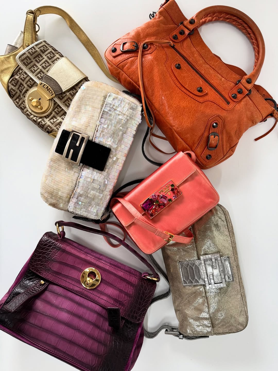 Six designer handbags in various colors and styles are arranged on a white surface, including metallic gold, woven white, coral, orange, deep purple, and silver bags with distinctive clasps and hardware.