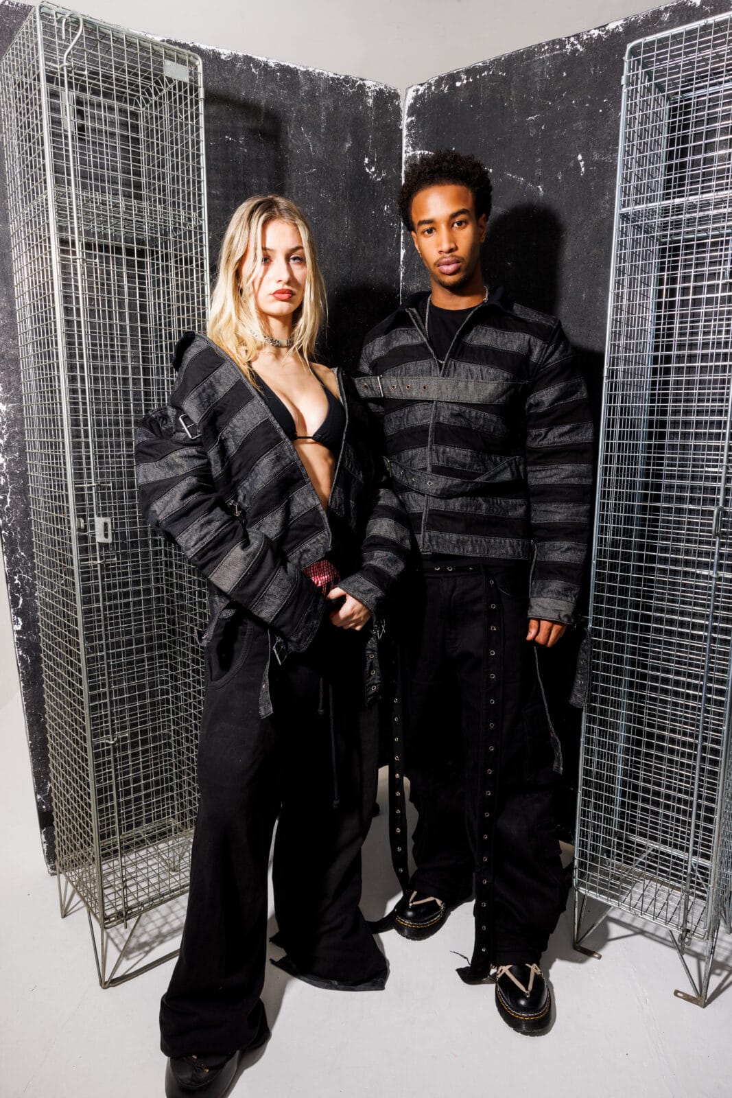 Two models stand side by side in front of wire lockers, wearing All-T¥lk coordinated black outfits with wide horizontal stripes and details. The woman’s jacket is open, revealing a black bra, and both display confident expressions.