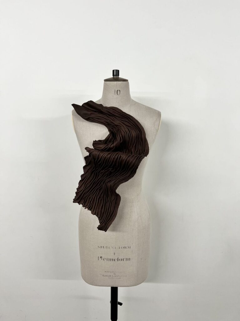 A Salma Tahir dress form displays a sculptural, asymmetrical brown garment with dramatic pleats cascading diagonally across the torso against a plain white background.