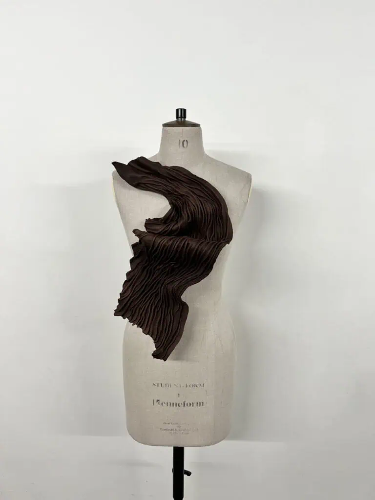 A Salma Tahir dress form displays a sculptural, asymmetrical brown garment with dramatic pleats cascading diagonally across the torso against a plain white background.