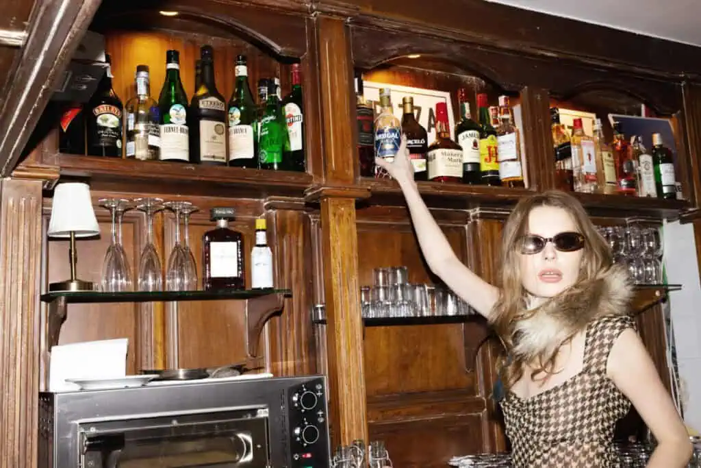 A woman wearing sunglasses and a fur-collared dress stands in front of a wooden bar, reaching for a bottle on the top shelf lined with various liquor bottles and glassware.