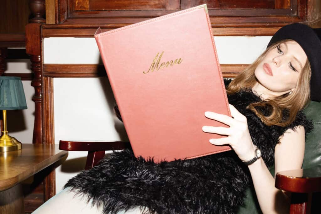 A woman in a black dress and beret sits in a chair, holding and reading a large menu. She appears relaxed and is seated in a warmly lit room with wooden decor in the background.