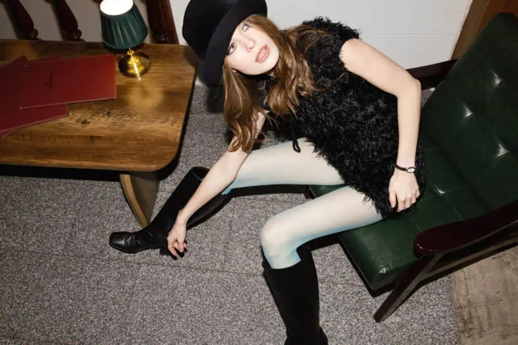 A woman in a black hat, black textured dress, white tights, and black boots sits on a green armchair, adjusting her boot. A wooden table with red folders and a lamp is beside her.