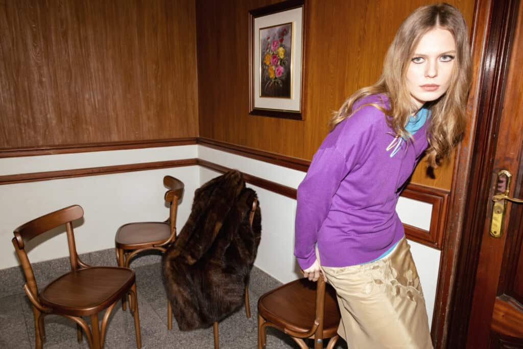 A woman with long hair wearing a purple top and beige skirt sits on a chair near a wooden door in a room with wood-paneled walls, three chairs, a fur coat, and a framed floral painting on the wall.