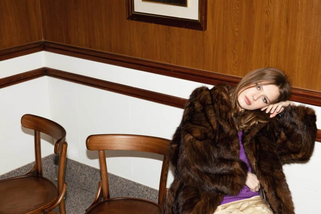 A woman wearing a dark fur coat and purple top sits alone on a wooden chair in a room with wood-paneled walls, resting her head on her hand and gazing pensively to the side. Two empty chairs are beside her.
