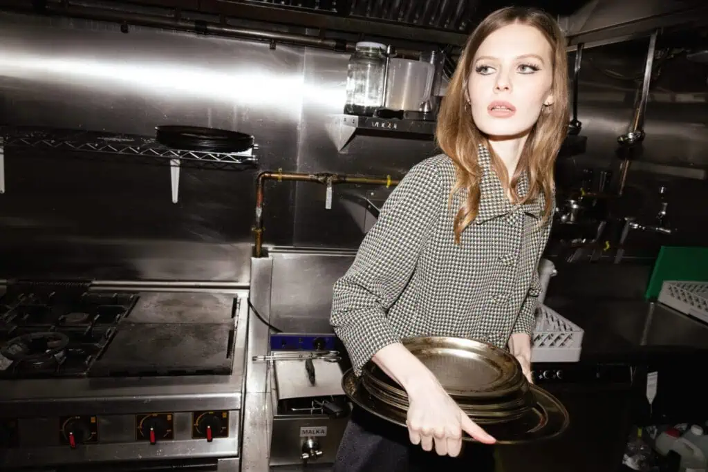 A woman in a checkered shirt holds several metal trays in a commercial kitchen, glancing to her right with a focused expression. Cooking equipment and shelves are visible in the background.