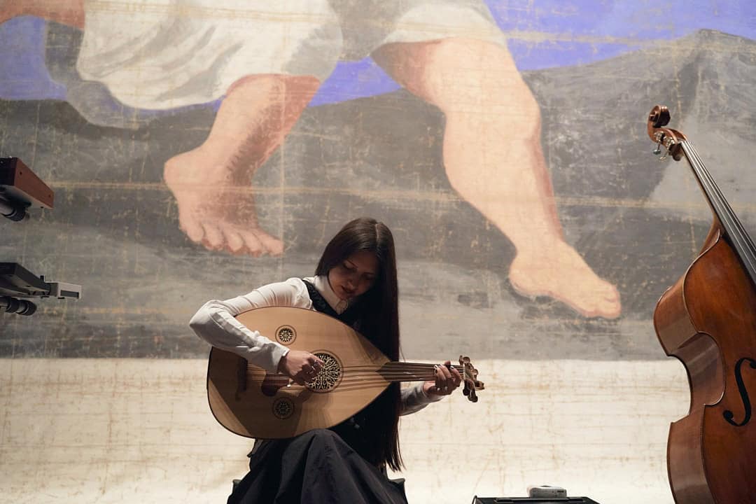 A wide shot of musician BODUR seated and playing a traditional oud. She is positioned in front of a large-scale mural, with a double bass visible to the right, highlighting her interdisciplinary musical style.