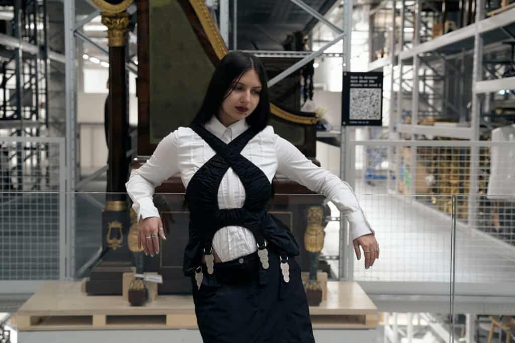 Portrait of musician BODUR wearing a black-and-white avant-garde outfit, leaning against a glass railing in a high-ceilinged industrial gallery.
