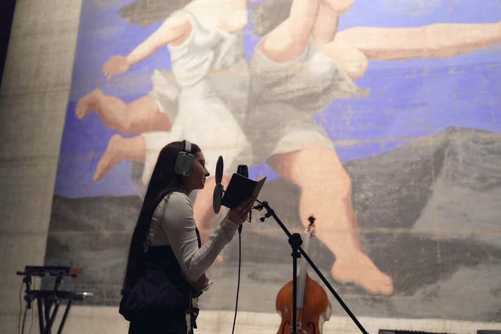 Side profile of BODUR singing into a studio microphone while wearing silver NOTHING(R) over-ear headphones, set against a large blue mural and a double bass.