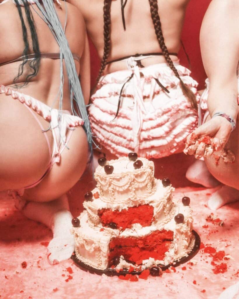 Three people in frilly, pink lingerie kneel on a pink furry surface around a partially eaten, three-tiered red velvet cake with white frosting and cherries, against a red background. One person is holding cake in their hand.