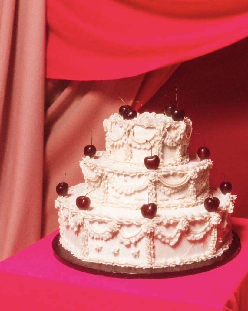 Three-tiered white cake decorated with ornate piped icing and topped with cherries, displayed on a bright pink surface with red and pink draped fabric in the background.
