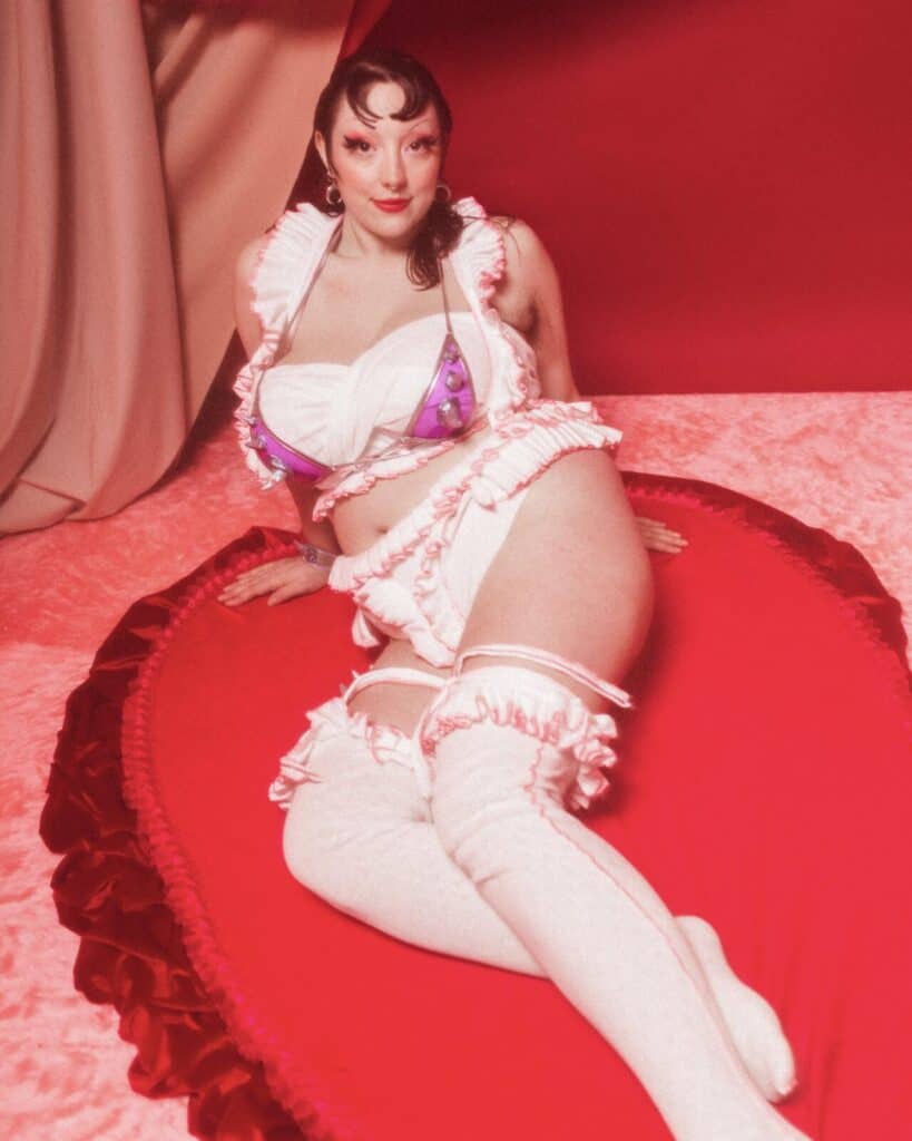 A person with vintage-style makeup and curled hair poses on a plush, red, heart-shaped cushion. They wear frilly white lingerie, white thigh-high stockings, large hoop earrings, and are surrounded by pink and red fabric.
