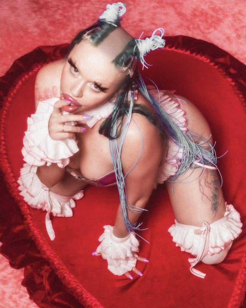 A person with unique hair buns and blue extensions poses provocatively on a red heart-shaped couch, wearing frilly white accessories and pastel makeup, set against a pink and red background.
