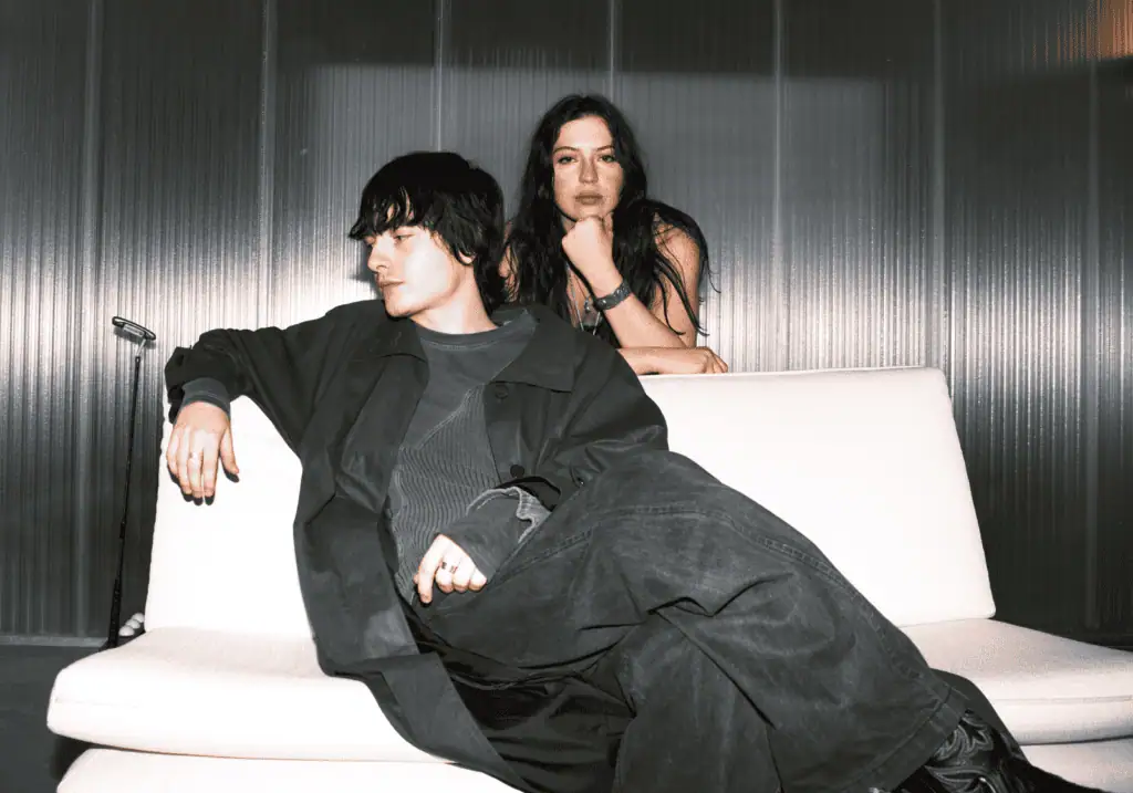 Witch Post band members posing on a white sofa in a dimly lit room, showcasing their dark, alternative fashion and indie rock aesthetic.