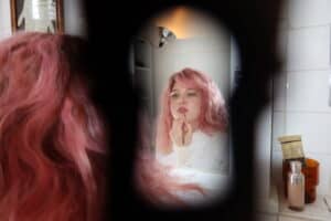 A creative shot of Grace Ives seen through a keyhole-like frame, showing her reflection in a bathroom mirror while she applies makeup.