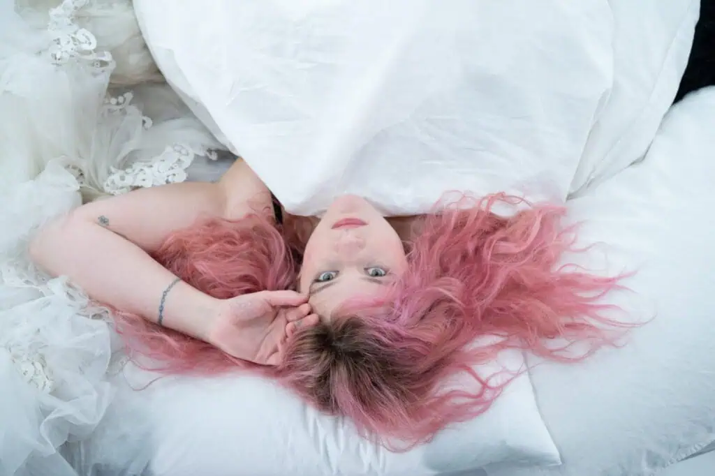 Grace Ives lying on white pillows with her long pink hair spread out, looking directly at the camera in a bright, ethereal setting.