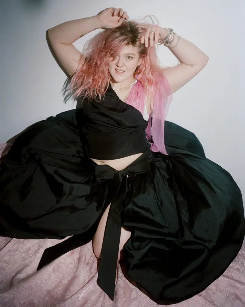 Grace Ives posing in a voluminous black balloon skirt and top, with her vibrant pink hair contrasting against a simple white background.