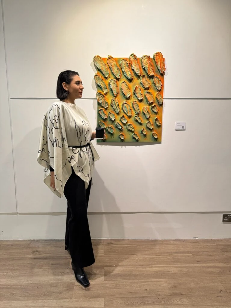 Halise Karakaya standing next to one of her highly textured orange and green impasto paintings during an art exhibition.