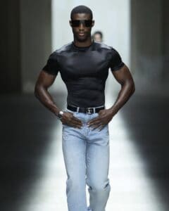 A male model walks for Gucci AW26 runway wearing a fitted black t-shirt, light blue jeans, dark sunglasses, and a black belt, with hands on his hips and a confident expression in a dimly lit setting.