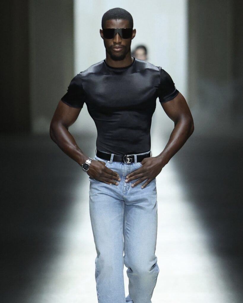 A male model walks a runway wearing a fitted black t-shirt, light blue jeans, dark sunglasses, and a black belt, with hands on his hips and a confident expression in a dimly lit setting.