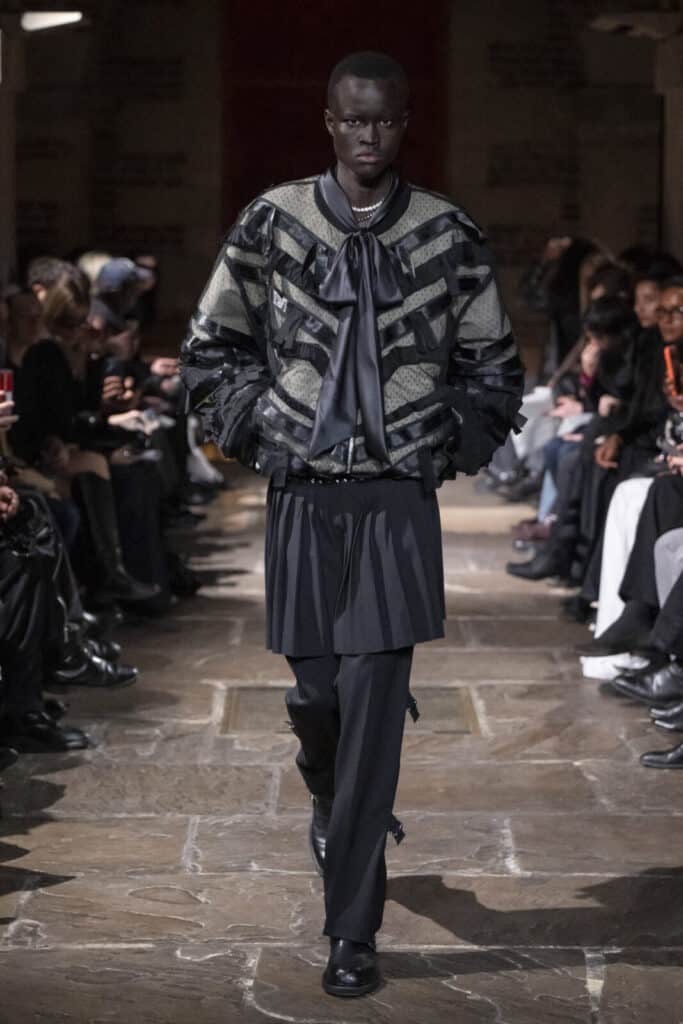 Model wearing a textured grey bomber jacket over a pleated kilt and trousers by John Richmond