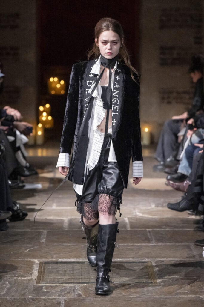 Model on the runway wearing a velvet blazer with "Angel" text and lace-trimmed shorts by John Richmond