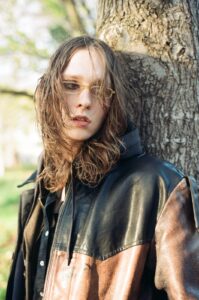 A portrait of Lauren Auder leaning against a large tree trunk, wearing a brown and black leather jacket and yellow-tinted glasses in soft natural light.