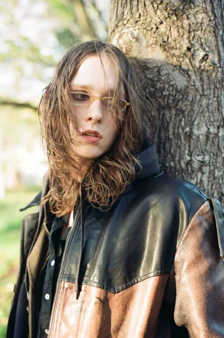 A portrait of Lauren Auder leaning against a large tree trunk, wearing a brown and black leather jacket and yellow-tinted glasses in soft natural light.