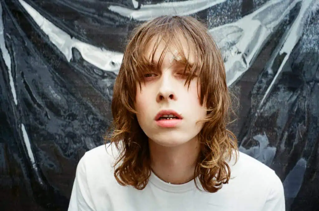 A close-up headshot of Lauren Auder with long hair and a white t-shirt, posed against a textured metallic silver background.